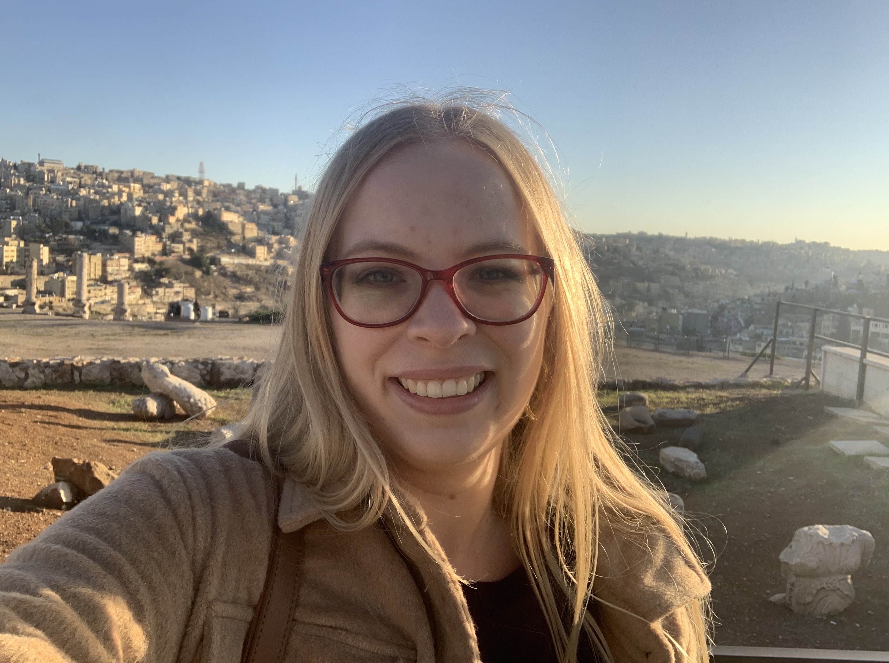 Photo of a young white woman with long, blond hair and red glasses, wearing a tan colored coat. The skyline of Amman, Jordan is visible in the background.